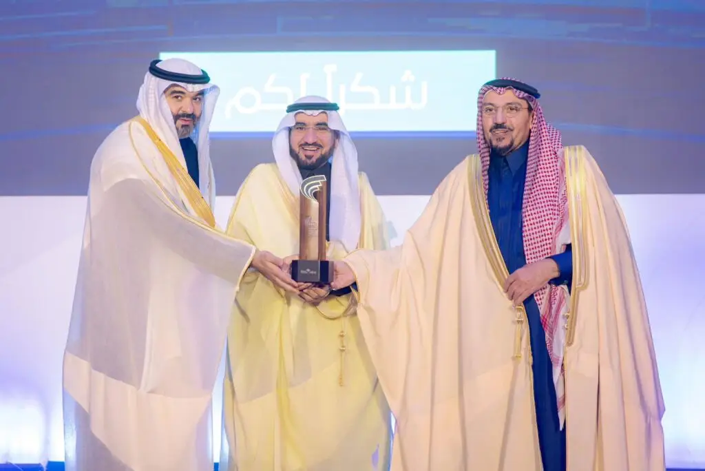 كل ثلاثة رجال تولي الزي السعودي الأصلي، يشملاً الشماغ أو الغترة السعودية، معاً على خشبة المسرح خلال ملتقى الأعمال تعهيد، وهم يبتسمون ويحملون درعاً تكريمياً. كتب في الخلفية لوحة مضيئة تحمل نصوصًا باللغة العربية تتضمن اسم الجامعة وأمير منطقة القصيم.
