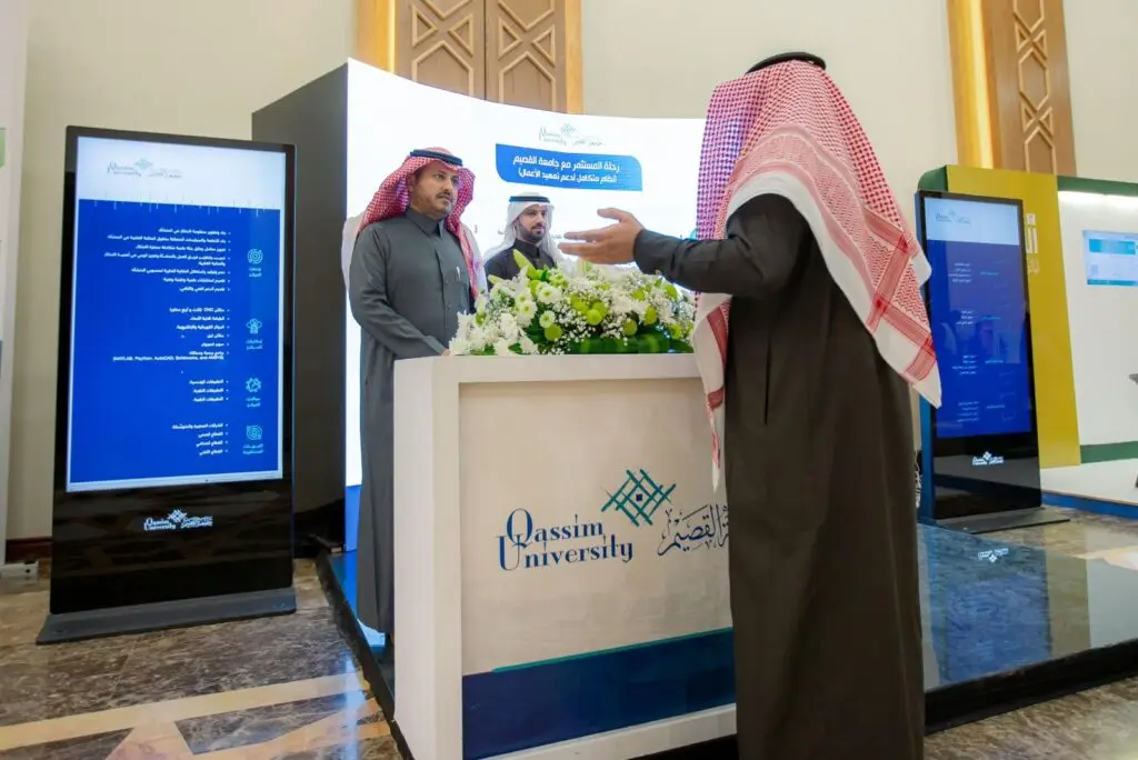 رجل يرتدي بدلة سعودية أصلية مع شماغ سعودي، وكبارًا إلى شخصيين في جناح المعلومات لجامعة القصيم خلال ملتقى تعهيد الأعمال، ويتميز الجناح بوجود برمجية وشاشات رقمية كبيرة تعرض معلومات عن الجامعة العربية.