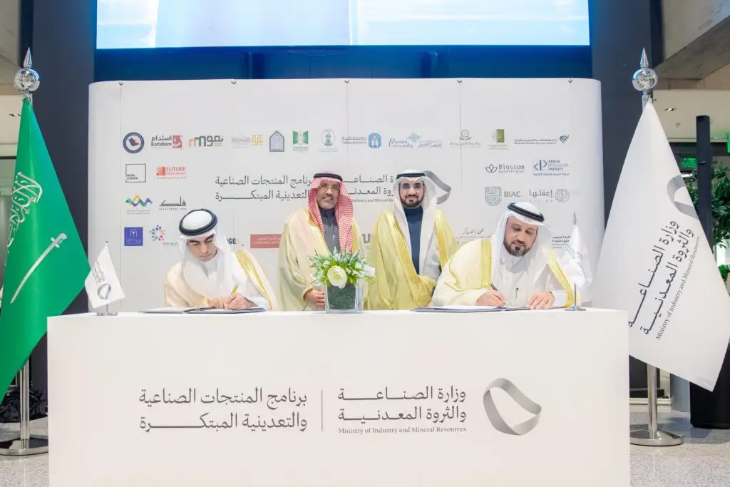 العروس تلتزم بالشماغ السعودي والثياب التقليدية عند طاولة بيضاء تحمل شعار وزارة الصناعة والثروة المعدنية، حيث تقوم بتوقيع المستندات اليومية وتؤكد التعاون في مجال الصناعة والتعدين. ويقف خلفهم آخران، بينما يبرز أعلام وشعارات الهيئات التنظيمية في، بما في ذلك تأكيد الحقيقة والرؤية المشتركة بين المشاركين.