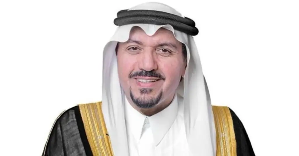 A man dressed in traditional Saudi garb - a white dress, black and gold bisht, and a white Saudi jacket tied with a black headband - stands smiling against a simple white background, embodying the pride and dignity associated with His Royal Highness the Emir of Qassim.