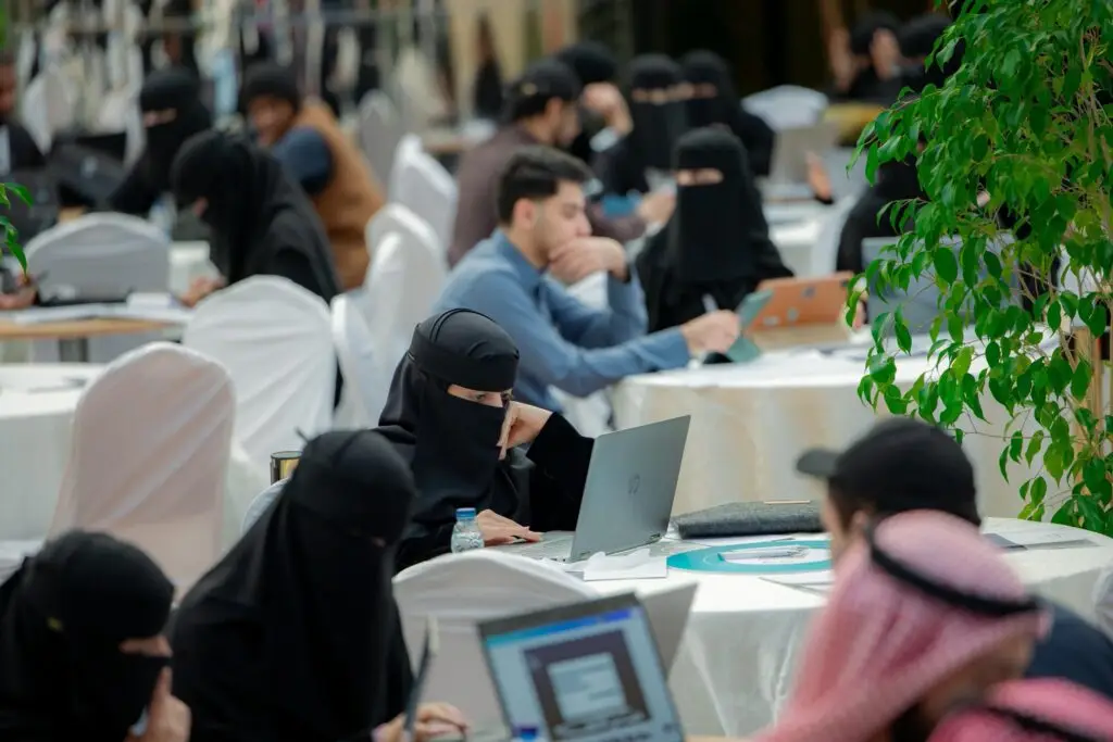مجموعة من الأشخاص، وعددهم من النساء يرتدين النقابات والعباءة السوداء، يعملن على أجهزة كمبيوتر محمولة حول طاولات مستديرة خلال هاكاثون حاضنة ابتكار في جامعة القصيم. وينضم إلينا بعض الرجال مرتدين الشماغ السعودي أو الغترة السعودية مع العقال والبشت السعودي أو الزي الرسمي أو غير الرسمي، فيتعين علينا أن نتوصل إلى تطوير مشاريع ابتكارية.