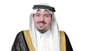 A man wearing a traditional Saudi dress, with a black Saudi Ghutrah with a black hood, a black jacket embroidered with gold on his shoulders, smiling in front of a crisp white background, is attending an official graduation at Qassim University under the patronage of His Highness the Emir of Qassim Region, and finally smiling in front of a crisp white background.