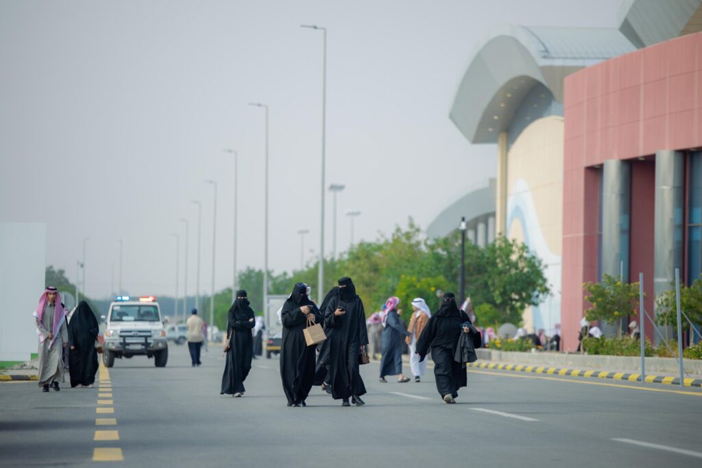 يسير عدد من الأشخاص على طريق أقل من مبنى حديث، حيث تقضي معظم النساء بعباءات ونقابات سوداء. أما بعض الرجال، والذين يُحتمل أن يسمى خريجو الجامعة، فيرتدون الزي السعودي التقليدي في الشماغ أو الغترة السعودية مع العقال والبشت السعودي. التوقف في الصورة الخلفية لمركبة أمنية بجوار المبنى.