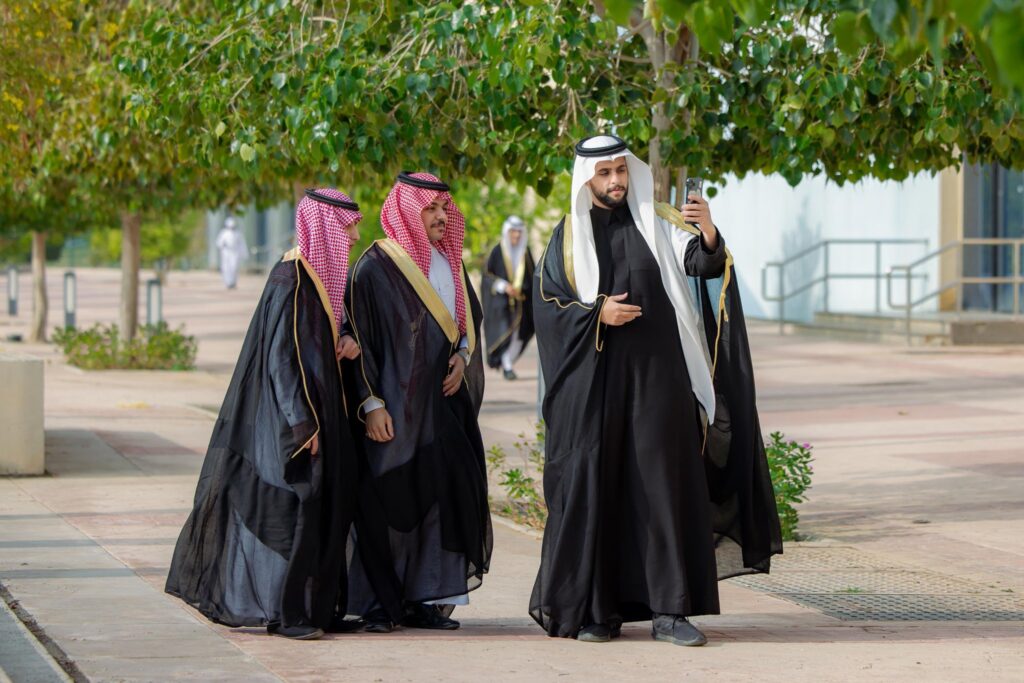 ثلاثة رجال كذبوا تقليدياً كما غترة سعودية أو شماغاً، مع قال وبشت سعودي، الاتصالون في الهواء الطلق في يوم مشمس الأثرن مبتسمين بينما يقوم أحد بالتقاط صورة ذاتية بهاتفه المحمول. ومن المحتمل أن يكون خريجون من جامعة القصيم، وتحيط بهم نسخ وممرات مرصوفة.