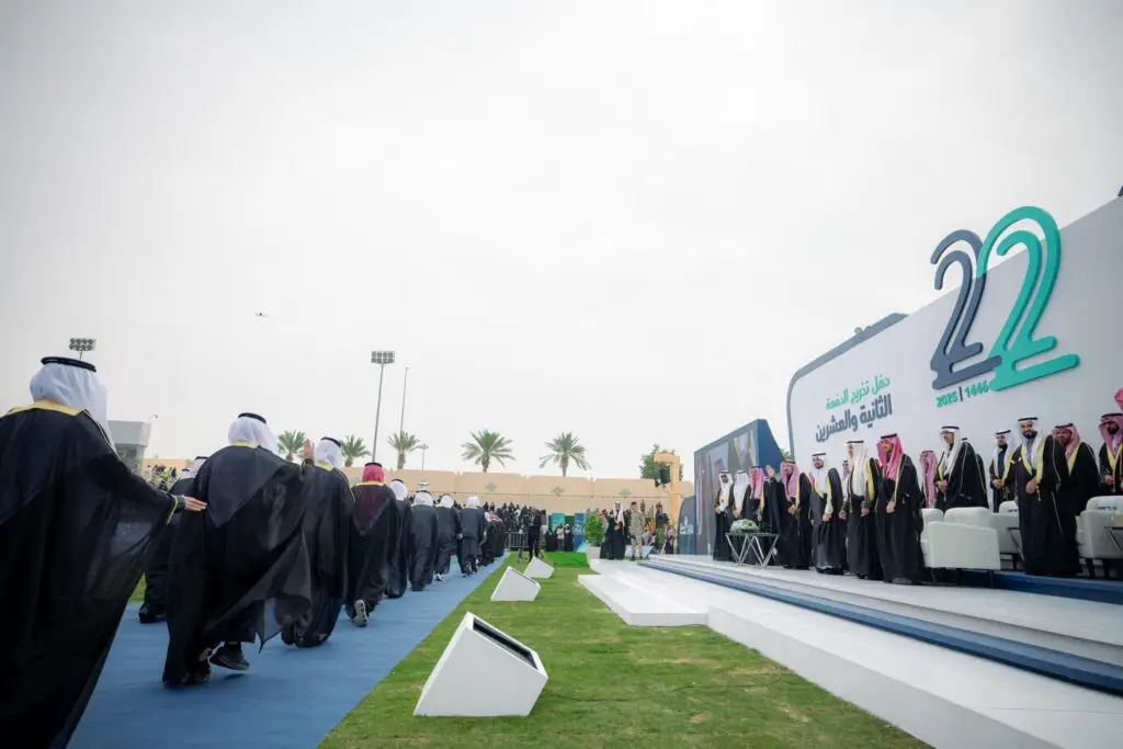 في الهواء الطلق، يشارك فيه الحضور الزي الرسمي السعودي، بما في ذلك الخريجون الذين سيتوجهون نحو المنصة ويجلسون معهم المسؤولون وأمير منطقة القصيم. تعلّم في الخلفية رقم 22 ونصوص عربية ذات صلة في القصيم ونخيل وسماء غائمة.