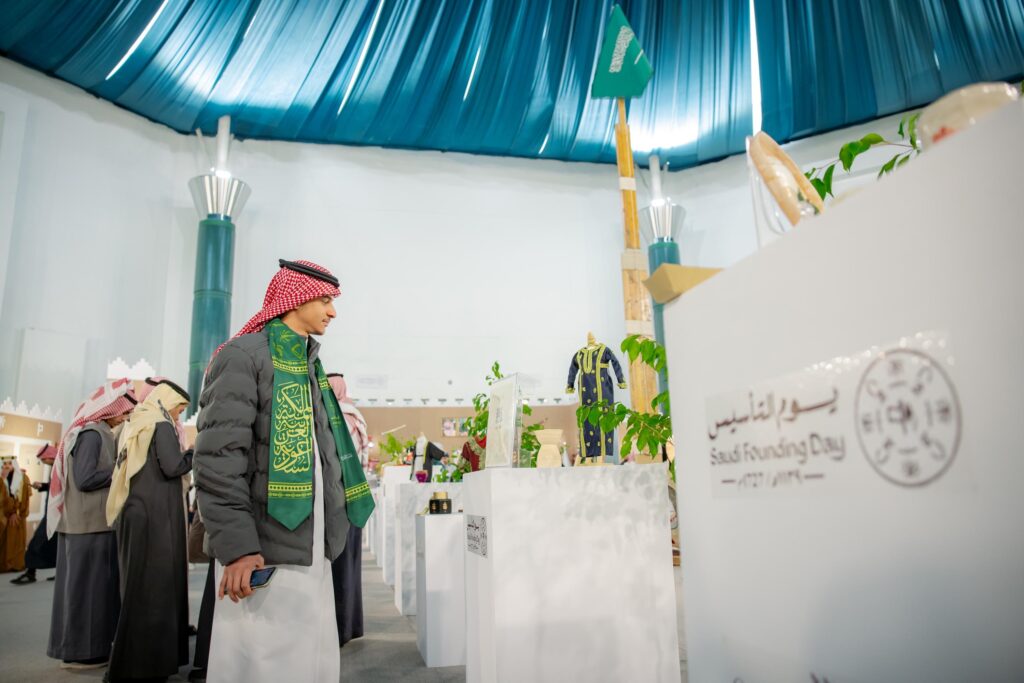 يقوم رجل يرتدي الغترة السعودية والعقال والبشت السعودي، بالإضافة إلى الوصية الخضراء، بتفقد معروضات في معرض الخلفية الداخلية يُقام في جامعة القصيم احتفاءً بيوم وتسعة السعودي، حيث يعيش في أعلام المملكة وبعض القطع التراثية.