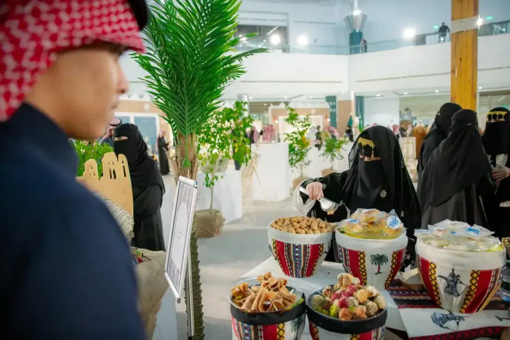 في سوق داخلي خلال فعاليات يوم التأسيس بجامعة القصيم، تُقدّم نساء يرتدين العباءات السوداء والنقاب حلويات شرق أوسطية تقليدية في أوعية ملونة. وعلى مقربة، يقف رجل يرتدي شماغًا سعوديًا بانتباه. ويزيد من حيوية المكان وجود النباتات الخضراء، مما يُضفي أجواءً نابضة بالحياة.