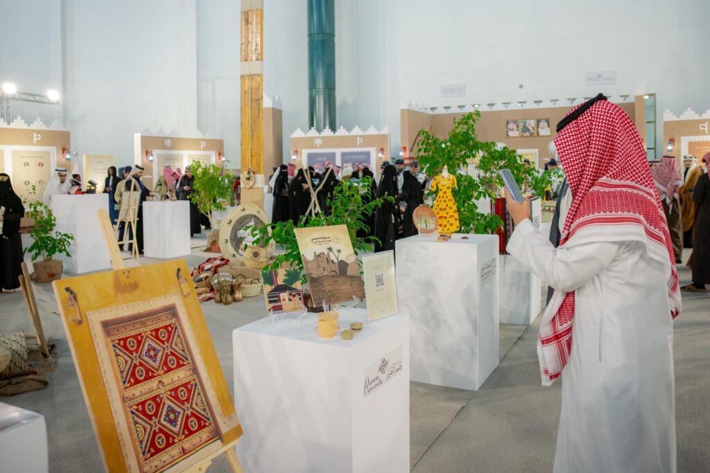 ارتدى الرجل الزي السعودي الرسمي، بما في ذلك الغترة والنباتات والبشت السعودي، يلتقط صورة بهاتفه المحمول من خلال المعرض الثقافي الداخلي استضافته جامعة القصيم، ولكن العمل عملياً وفنية لا ينضب من الأخضر، بينما يظهر في الخلفية عدد من الزوار الآخرين.