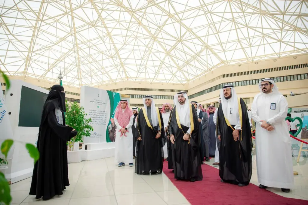 أتقابل مجموعة من الرجال مرتدين الشماغ أو الغترة السعودية السعودية، مع جانب فتاة ترتدي ملابس نسائية، على سجادة الحمراء داخل مبنى ذي سقف زجاجي في جامعة القصيم، وذلك من خلال تُبرز رمزية يوم العلم.