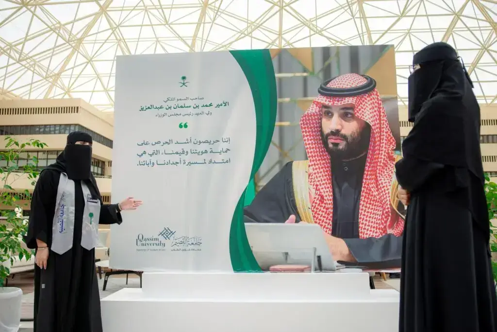 تتقف أمرأتان ترتديان ملابس بيضاء ونقاباً بجوار لوحة كبيرة لصاحب السمو الملكي الأمير محمد بن سلمان بن عبد العزيز آل سعود، تتضمن نصوصاً عربية وشعارات شعبية، وذلك في مساحة لا تتجاوز سقف زجاجي داخل الحرم الجامعي، مما يبرز مكانة العلم وروح يوم العلم في جامعة القصيم.