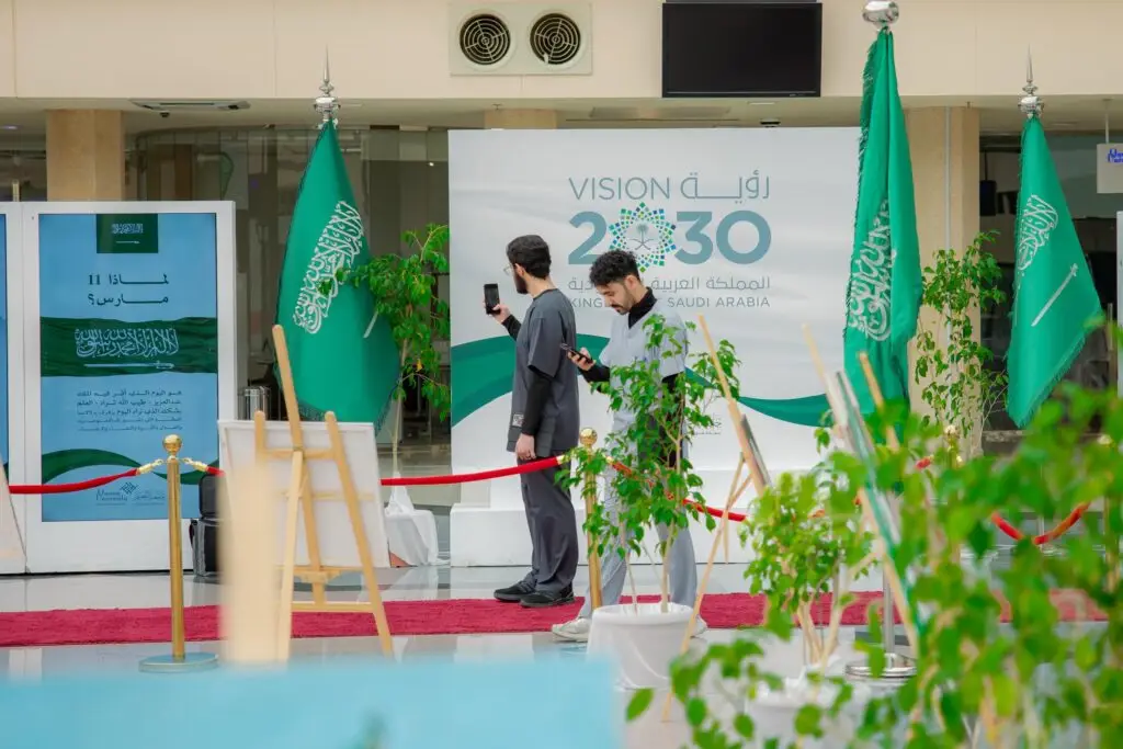 رجل الاتصال بالقرب من أعلام المملكة العربية السعودية ولوحة رؤية 2030 داخل إحدى الغرف، حيث يوجد المنطقة حواجز حمراء. احتفي بالمشروبات العشبية الخضراء في نهاية المطاف عرض توعوية تسلط الضوء على مكان يوم العلم، بينما يرتدي أحد الرجال شماغاً سعودياً تأكد من غترة سعودية، في مشهد يعكس بوضوح تليق في القصيم.