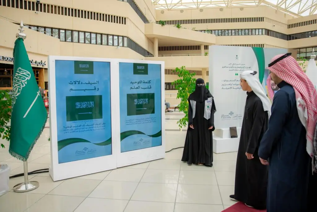 مجموعة من الأشخاص تولي الزي السعودي الأصلي، بما في ذلك الشماغ أو الغترة السعودية، يتابعون عرض النص الرقمي يحمل نصوصًا باللغة العربية من خلال فعالية احتفاءً بـ "يوم العلم"، وشرب راية المملكة العربية السعودية بناءً على ترخيصها. يتميز بالمكان بطرازه المعماري العصري ووفرة الضوء الطبيعي، وذلك ضمن نطاق التعليم في جامعة القصيم.