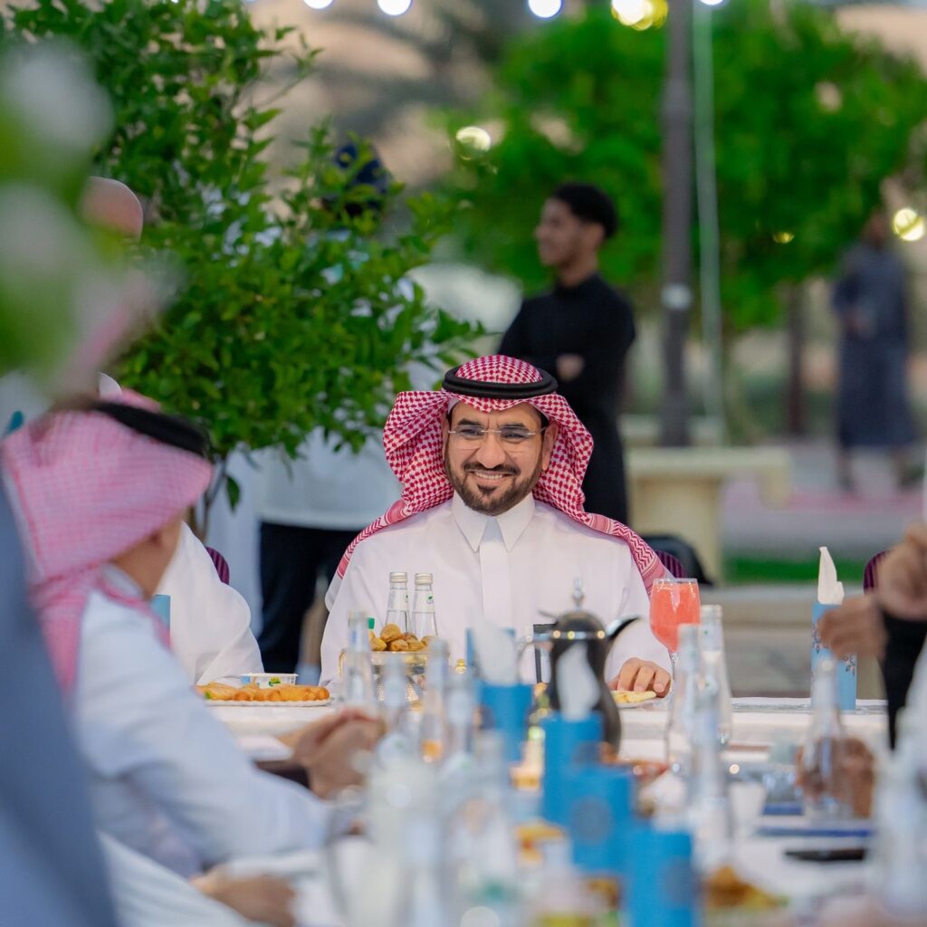 ارتدى رجل الغترة والعقال والبشت السعودي يجلس مبتسمًا على طاولة خارجية معدودة بالأطعمة إلا في جامعة القصيم، واجتماع جواره مجموعة من الأشخاص الشماغ والعقال والبشت السعودي أيضًا، بالإضافة إلى مساحة خضراء.