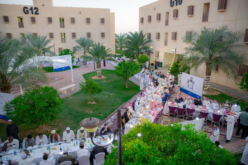 تجمع كبير في الهواء الطلق في القصيم، حيث يجلسون على طاولات طويلة في وسط الساحة، مع شركة النخيل والمباني الموسومة برمزي G10 وG12. ويظهر المشاركون وهم يحضرون الغترة أو الشماغ السعودي مع العقال، بالإضافة إلى البشت السعودي، في السيدات احترام الثقة الشخصية التي يُتوقع أن تكون أدبية جماعية أو فعالية تحت رعاية الجامعة.