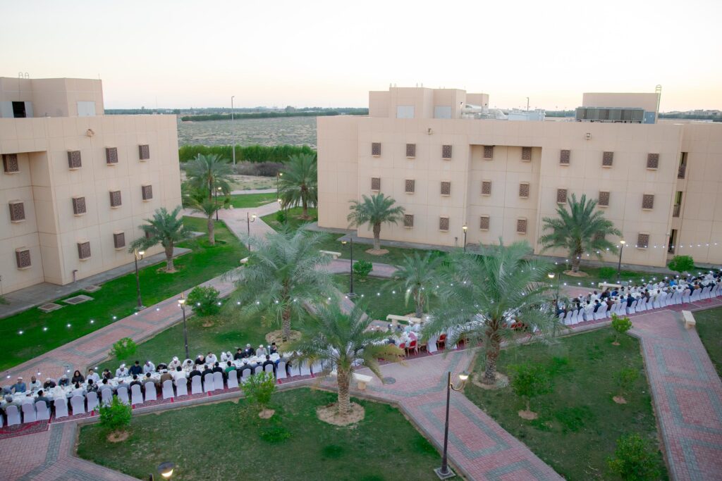 يصطف صف طويل من الأشخاص مرتدين الغترة أو الشماغ والعقال والبشت السعودي على امتداد موائد متتابعة الطبقة على طول ممر بين المباني في جامعة القصيم، وسط نخيل ومساحات خضراء أثناء تجمع خارجي يُقام قبيل غروب الشمس.