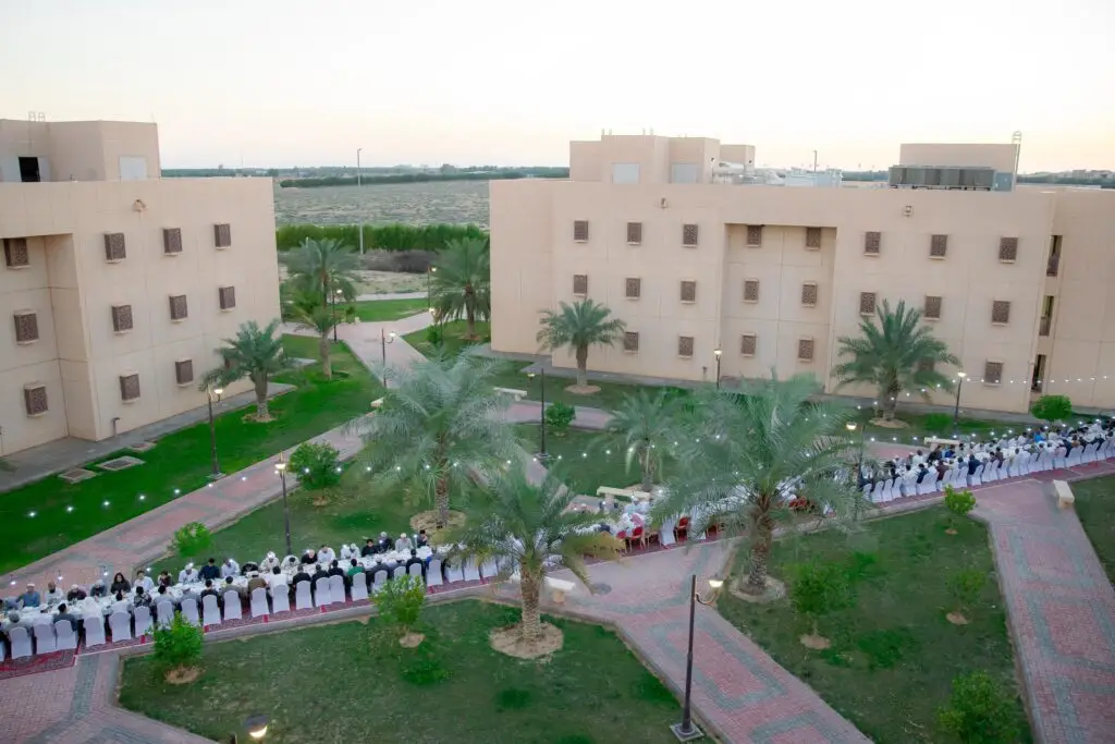 يجتمع جمع كبير من الناس، الفستان الأبيض التقليدي مع الشماغ أو الغترة السعودية، حيث تجتمع مائدة طويلة في الهواء الطلق بين المباني التجارية وأشجار النخيل. يأتي ذلك ضمن فعالية جماعية لتناول وجبة الإفطار عند غروب الشمس، وذلك في ختام برنامج الريان الرمضاني المتنوع من قبل جامعة القصيم.