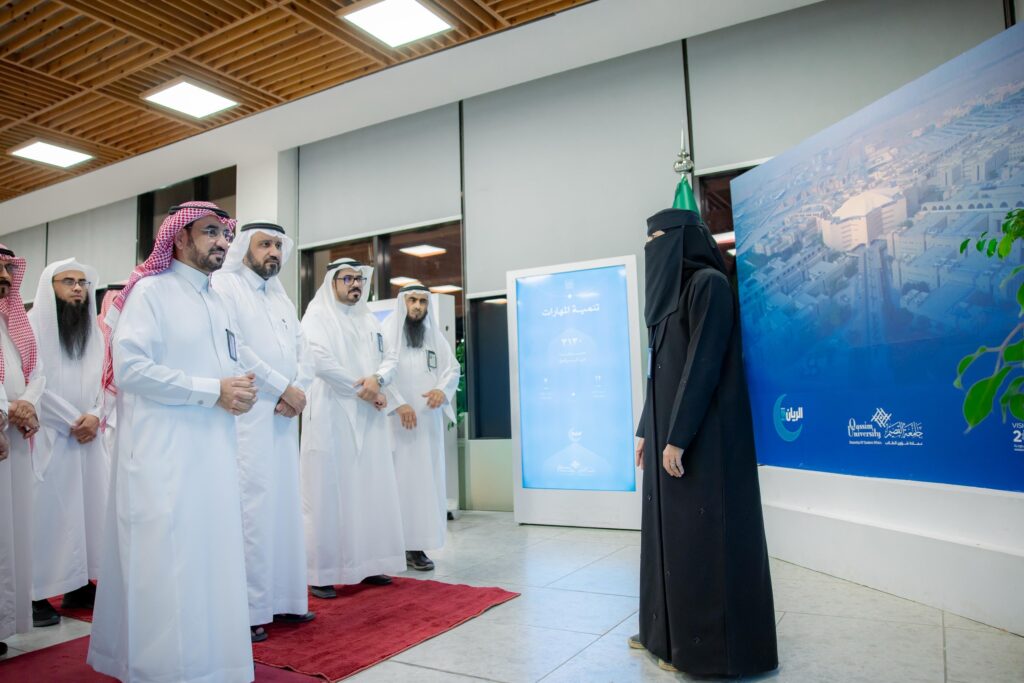 اتصلت مجموعة من الرجال مرتدين الثياب البيضاء مع الشماغات والعُقَل والبشوت السعودية، إلى جانب جانبهم ترتدي ملابس نسائية بيضاء ونقاباً، وذلك في صالة مفروشة بالسجاد الأحمر خلال ختام برنامج الريان الرمضاني، بحضور رئيس جامعة القصيم. خذ خلفهم شاشة عرض نصوصاً زرقاء تحمل نصوصاً وصوراً باللغة العربية.