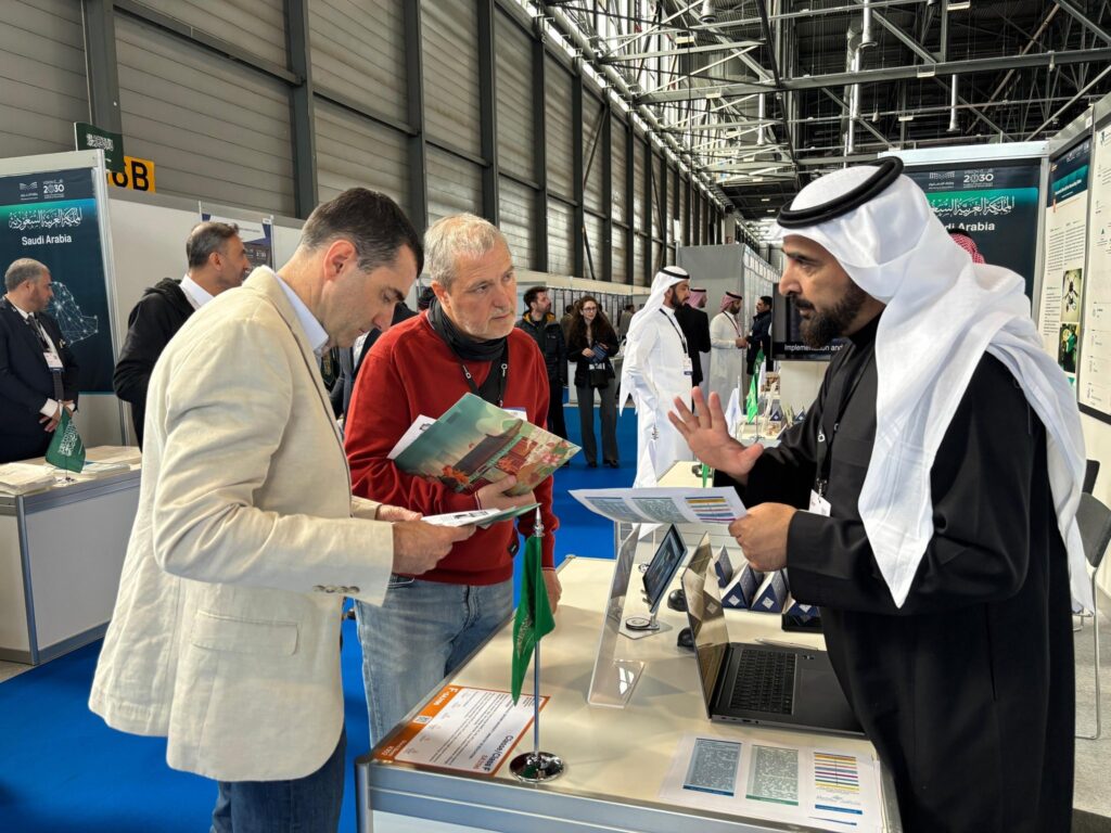 رجل يرتدي الغترة والعقال والبشتاء السعودي الكبير مع رجل أمام جناح جامعة القصيم في أحد المعارضين، بينما عصر الرجلان فوجات تعريفية ويصغيان باهتمام. وفي هذه الخلفية يظهر عدد من الزوار والأجنحة الأخرى، مما يجعلهم معارضين للمعارضة.