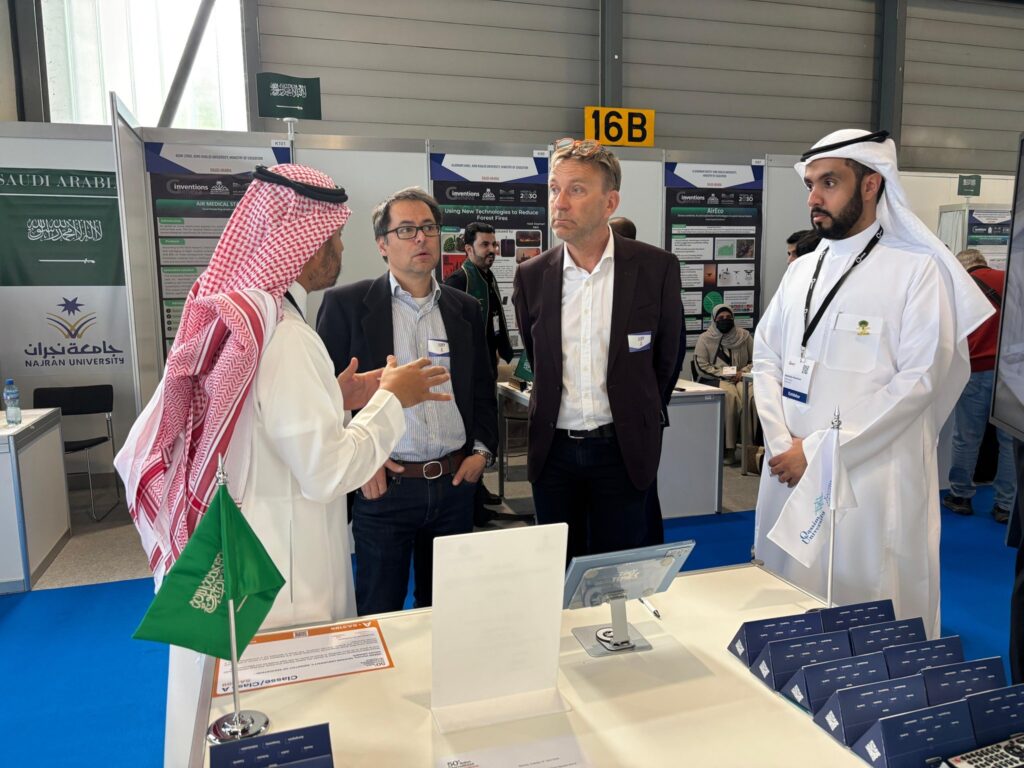 يتبادل أربعة رجال حديثين في جناح داخل قاعة المعرض؛ ارتدى اثنان منهم بزات غربية، بينما يظهر العشرات بزي سعودي عادة تشمل شماغًا سعوديًا أو غترة سعودية مع عقال وبشت سعودي. وتبرز في الجدول المطويات الرسمية للمنشورات جامعة القصيم، بالإضافة إلى أعلام المملكة العربية السعودية، في مشهد يعكس بوضوح المنافسة الرسمية للجناح.