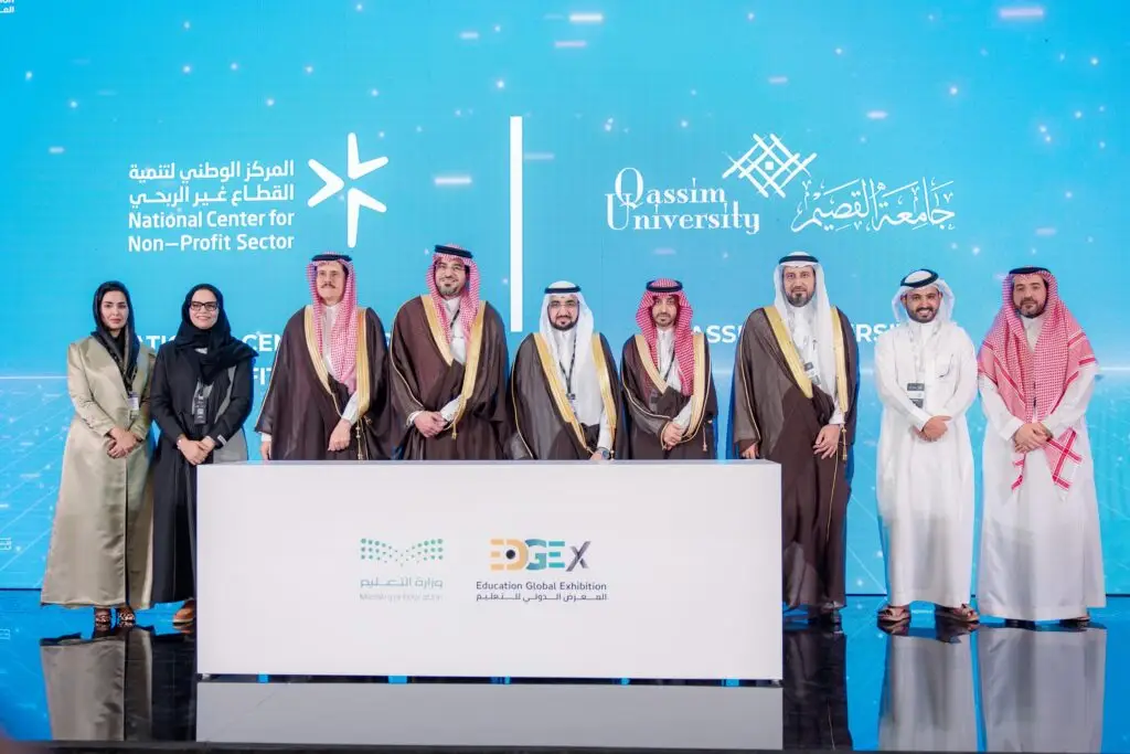 A group of men in a Saudi ghutra or shemagh with a Saudi headband, along with women in formal business attire, touch the stage in front of a large screen bearing the logos of the National Center for Nonprofit Sector Support and Qassim University, celebrating the efforts of the nonprofit sector and developmental studies.