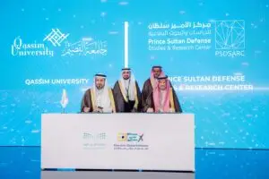 الرجال تولى الغترة والشماع والعقال والبشت السعودي، يوقعون وثائق أربعة أطفال على الطاولة، في مشهد يجسد التعاون البحثي في المجال الدفاعي الجديد. وخلفهم شعار جامعة القصيم ومركز الأمير سلطان للدراسات والأبحاث الدفاعية على خلفية رقمية زرقاء.