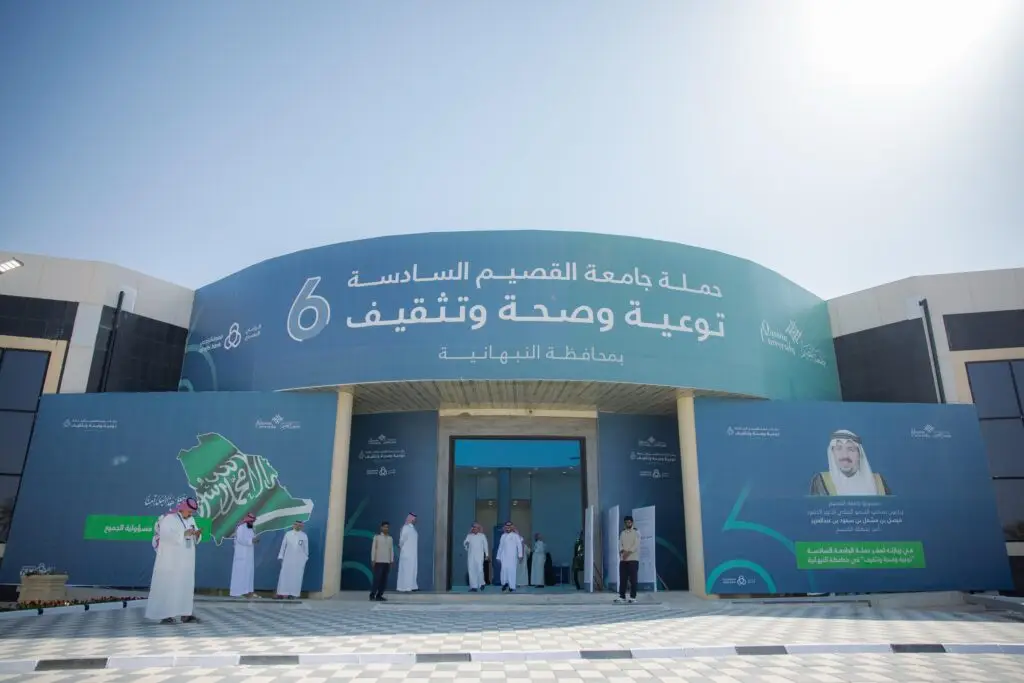 واجهة مبنى كبير أزرق يحمل نصوصاً وشعارات باللغة العربية، ويتواجد أمامها مجموعة من الأشخاص الزي السعودي الرسمي، من شماغ سعودي أو غترة سعودية مع عقال وبشت سعودي. وقفت لافتة تحمل علم المملكة وصورة الشخصيات المميزة، في إطار حملة توعوية وصحية تنظمها جامعة القصيم.