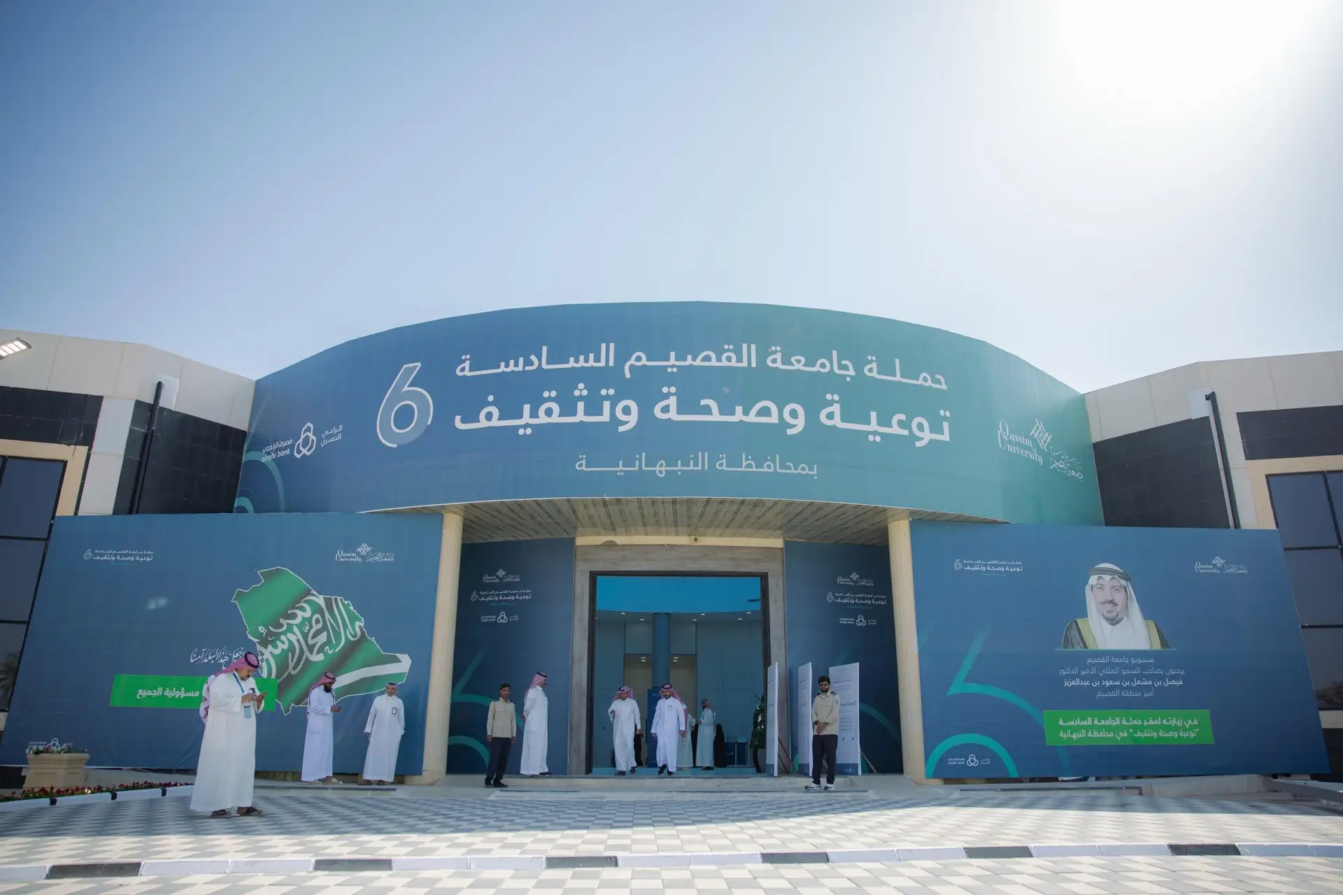 واجهة مبنى كبير أزرق يحمل نصوصاً وشعارات باللغة العربية، ويتواجد أمامها مجموعة من الأشخاص الزي السعودي الرسمي، من شماغ سعودي أو غترة سعودية مع عقال وبشت سعودي. وقفت لافتة تحمل علم المملكة وصورة الشخصيات المميزة، في إطار حملة توعوية وصحية تنظمها جامعة القصيم.