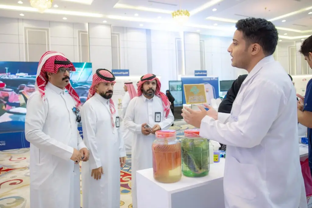 رجل يرتدي معطفاً أبيض في معرض حديث ومشرق، يشرح لثلاثة رجال يرتدي البشت السعودي والسترة والشماغ والعقال. تحتوي الضوء على أهمية التوعية الصحية، في حين تتواصلون جميعاً بجانب طاولاتها تجربان كبيرتان سوائل، وذلك ضمن فعاليات جامعة القصيم.