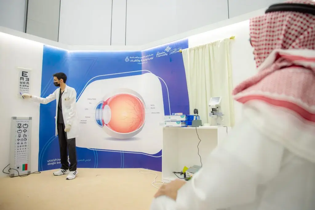يقوم أحد المختصين في الرعاية الصحية، ارتدياً المعطف الأبيض والكمامة الطبية، بالإشارة إلى لوحة فحص النظر بينما يشرح أحد متقدمين الجالسين والذي يرتدي الغترة السعودية مع العقال والبشت السعودي. وفيها لوحة زرقاء تحمل شعار جامعة القصيم وتبرز كلمات: توعية وصحة وتثقيف العين.