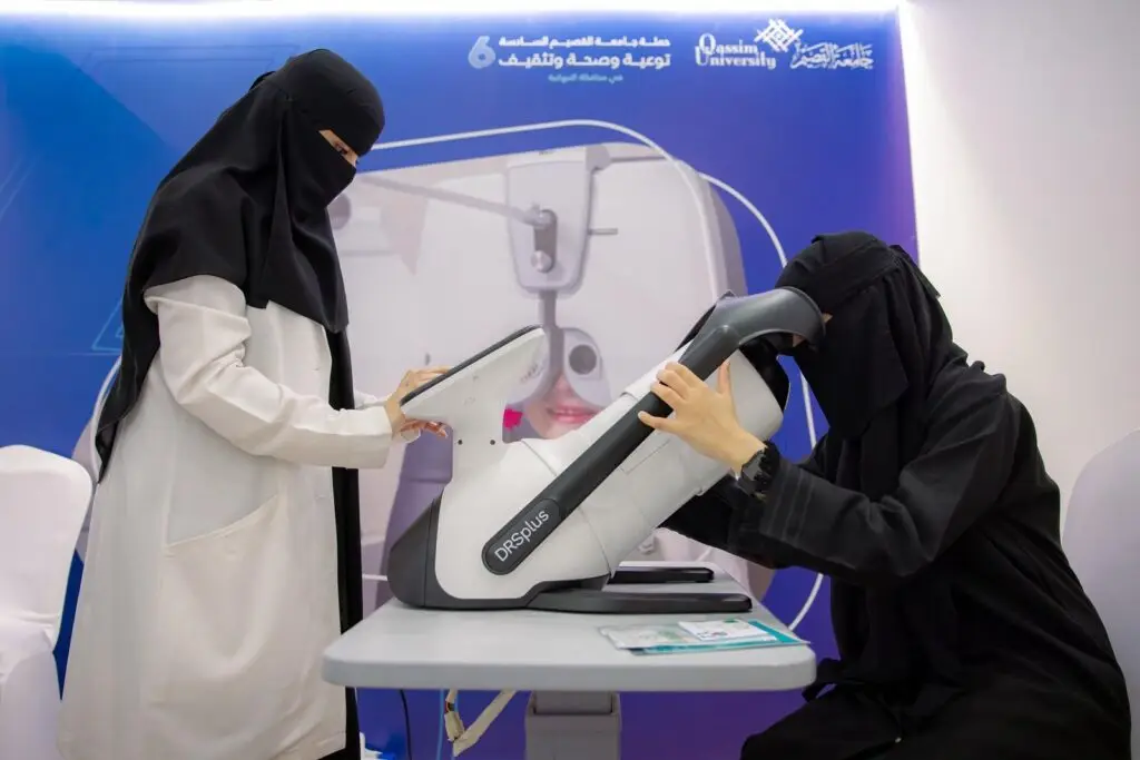 تمارس امرأتان ترتديان العباية والبشت السعودي مع الغترة والعقال، وهما تجريان فحصًا طبيًا للعين باستخدام خبير في إحدى العيادات. ويبرز في خلفية المشهد لوحة توعية باللون الأزرق تحمل نصوصًا باللغة العربية حول موضوع الصحة، بالإضافة إلى شعار جامعة القصيم، مما يسلط الضوء على جهود الرعاية الصحية المقدمة في مقاطعة النبهانية.