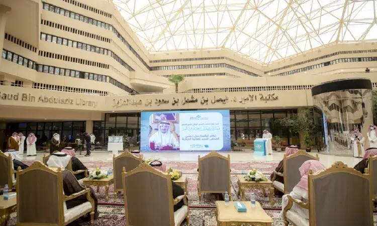 An official event is being held at the Qassim University Library inside a vast, progressive atrium, and after passing through its windows and architectural ceiling, a large screen displays an Arabic text and a portrait of His Highness Prince Dr. Faisal bin Meshal. A large screen bears Arabic text and a picture of His Highness Prince Dr. Faisal bin Meshal. Signs of respect for the library's national culture stand out, while some of the attendees are dressed in the Saudi Ghutra or Shemagh, Egal and Bisht, conveying the formality and atmosphere of the prestigious university.