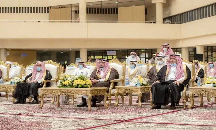 His Highness Prince Dr. Faisal bin Mishaal sits among a group of men wearing the Saudi Ghutra or Shemagh with Egal, and wearing the traditional Saudi Bisht, on detailed chairs with gold details, surrounded by beautiful olive flowers. This came during an event for the first time in a spacious hall with a luxurious carpet at the Qassim University Library, in the presence of a crowd of attendees.
