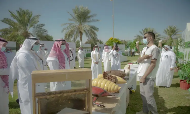 A group of men in Saudi Arabian ghutra or shemagh shirts, with Saudi Arabian aqal and bisht, listen to the conversation of a participant wearing a beige shirt, at an outdoor exhibition where wine, palms and plants are displayed on tables. This comes as part of the efforts of Qassim University's "Qassim Green Earth" campaign to plant 100,000 trees amid palm trees, pashtas and green spaces.