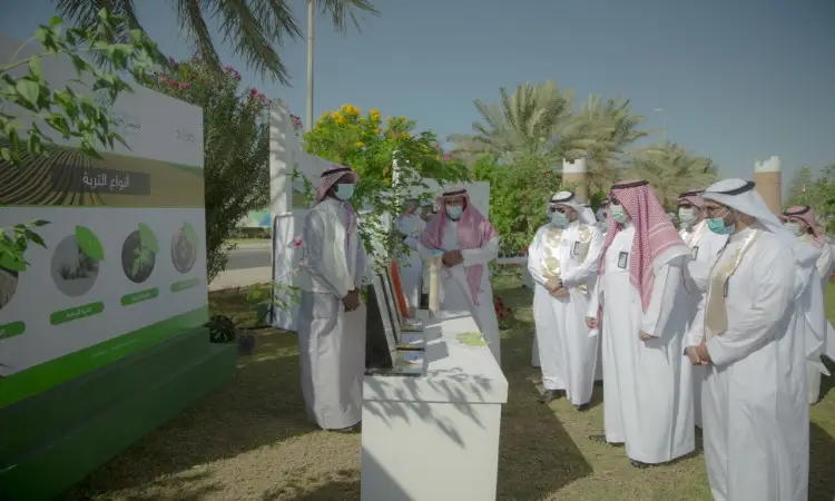 A number of men dressed in ghutra or shemagh and aqal, in addition to the Saudi bisht, gather in a green outdoor area near a display table and an informational board, partnering with them to collaborate and plant palm trees. This highlights the green "plant it" initiatives in the land of Qassim, which aims to plant 100,000 trees under a clear blue sky, as part of an effort sponsored by Qassim University to promote green agricultural sustainability.