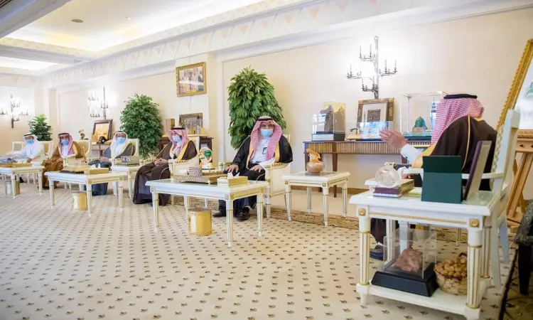 يجلس عددٌ من الرجال مرتدين الغترة أو الشماغ السعودي مع العقال والبشت السعودي على الكراسي، متباعدين في قاعة فخمة ومضيئة داخل جامعة القصيم. يحيط بالمكان لوحات فنية إبداعية ونباتات وثريات رائعة، بينما توزع أمامهم جداول متعددة الجمال.
