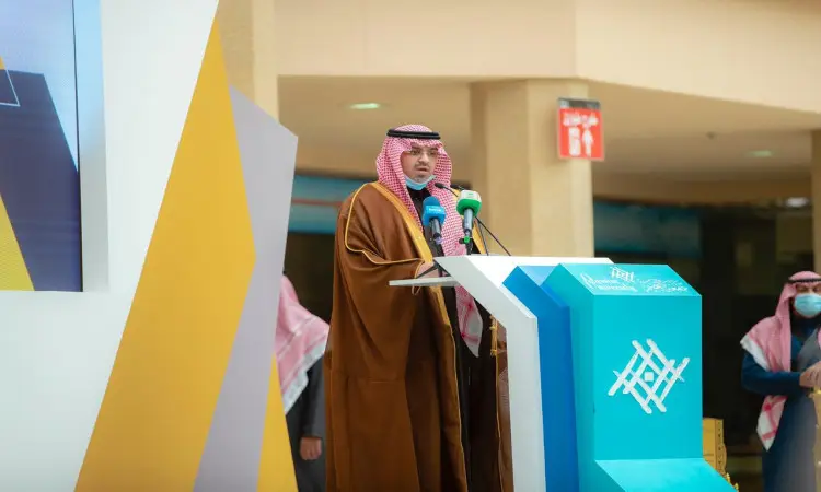 The man, often wearing a Saudi shemagh, headband and bisht, is standing at a podium with microphones, adorned with a blue-red banner. The event is held at Qassim University to highlight the achievements and developments taking place in Saudi Arabia, with a number of attendees in the background wearing the Ghutra or Shemagh and Egal in particular.
