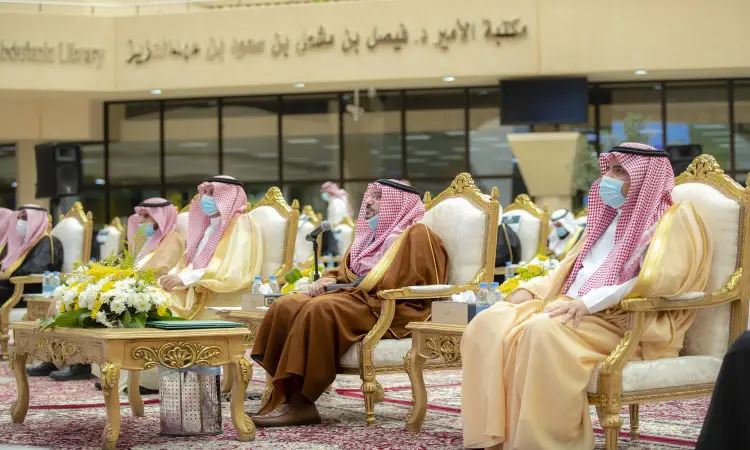 A number of men dressed in ghutra or shemagh and aqqal, as well as the Saudi bisht, sit on plush chairs during a children's event in Qassim held in a hall decorated with floral interiors and a plush carpet; behind them are books in Arabic pinned to the wall.