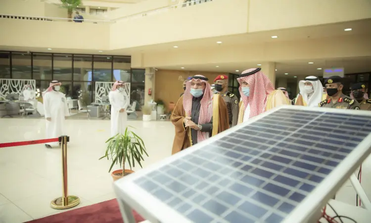 A group of men wearing Saudi Arabia's Ghutra or Shemagh, Egal and Bashtet, along with officers in their military uniforms, connect in a modern setting next to a military solar display. The scene is shown at Qassim University in Saudi Arabia, amid modern décor, plants, and a number of attendees dressed in formal white clothes.