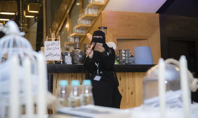 تجلس المرأة ترتدي سترة، وتضع غترة سعودية رسمية مع عقال على رأسها، فوق مقعد في أحد المقاهي، ممسكة بوريقة، ضمن فعالية يوم الميكروفون العربية فقط من جامعة القصيم. لكتابة خلفها طاولة خشبية تزينها أوعية وحاويات أنيقة؛ بينما تتغذى على مخلفات القش المزخرفة وقفص أبيض للزينة.