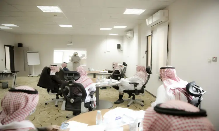A group of men dressed in ghutra or shemagh and aqal, as well as the Saudi bisht, gather around tables in a conference hall in Qassim, listening intently to a panelist's presentation on talent leadership.