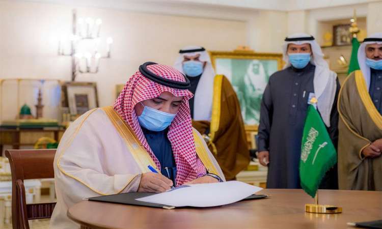 رجل ينتظر الغترة السعودية مع العقال والبشت السعودي وثوباً رسمياً، واضعاً كمامة طبية، وثيقةً على طاولة تتوسطها راية المملكة العربية السعودية؛ آثار ذلك ضمن إطار التوقيع بين جامعة القصيم ومستشفى الملك فيصل. الاتصال خلفه ثلاثة رجال وجبات بلباس رسمية تمثل الشماغ والعقال والبشت السعودي، جميعهم ملتزمون بارتداء الكمامات الطبية، وذلك داخل قاعة فخمة ذات طابع مميز يعكس عراقة المكان.