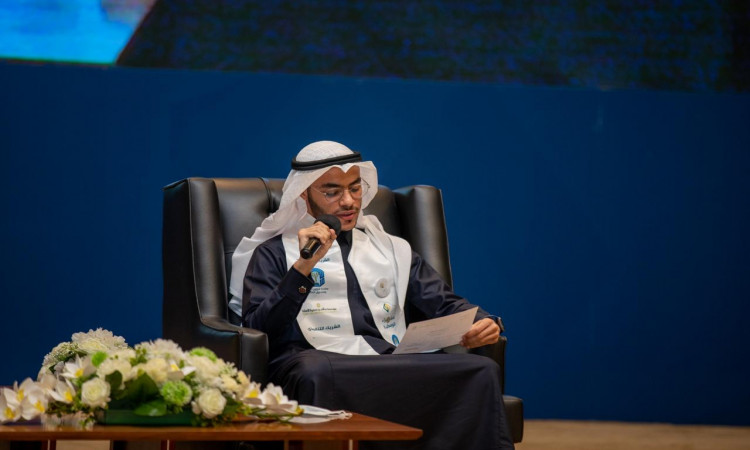 يجلس الرجل مرتدياً الغترة والعقال والبشت السعودي على مقعد أسود، ويضع شماعاً أبيض على كتفه، ممسكاً بيده تلقائياً أثناء قراءته من ورقة أمامه. وتزين طاولة أمامه بزهرة الزهور، بينما يظهر في الخلفية شعار جامعة القصيم في مدينة بريدة بلون أزرق رسمي.