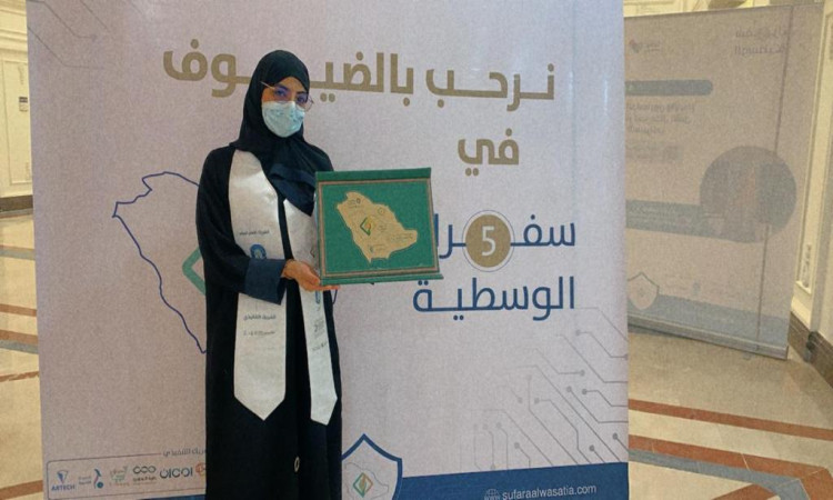 في مكان في القصيم، في الوقت الذي ترتدي فيه غترة سعودية وكمامة سيدة وبشتاً سعودياً وتحمل بيدها درع التكريم، وذلك أمام لوحة عرض تتضمن نصوصاً باللغة العربية وخريطة ثم، ممثلةً لمبادرة "سفراء الوسطية" في الجامعة.