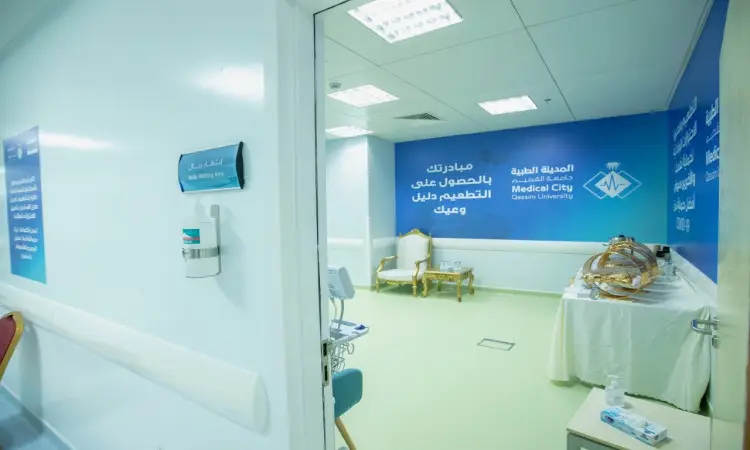طبية نظيفة ومضيئة بجدران خضراء في غرفة مستشفى جامعة القصيم، أبرزت لافتة خاصة باللغتين العربية والإنجليزية تشير بوضوح إلى المركز لتطعيم فيروس كورونا. تحتوي على غرفة جلوس وطاولة تشعر بغطاء أبيض مُعدة للاستشارات الطبية، بالإضافة إلى الأجهزة الطبية اللازمة. نتائج البحث في وجود بعض المراجعين أو الكادر الطبي مرتدين الشماغ السعودي والعقال أو الغترة مع البشت الرسمي السعودي، بما في ذلك بما في ذلك الهوية الوطنية والمظهر الرسمي الملائم لمكانة جامعة القصيم كمؤسسة أكاديمية موثوقة مرموق.