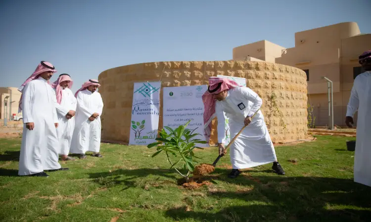 A small group of men take on the Saudi Arabian tunic or shemagh, aqal and besht, while someone plants a sapling in a tea area near circular buildings and banners bearing Qassim University's slogans about environmentalism and green initiatives.
