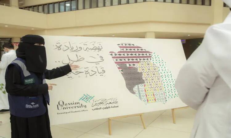 She covers her face with a black niqab and wears a traditional shemagh. In modern times, she is seen in front of a large canvas featuring Arabic calligraphy and colorful geometry. Standing near her are a number of participants inside the hall on the anniversary of the sale.