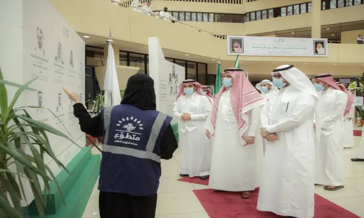 Volunteers explain information displayed on an information board about the university's exhibition to a group of men dressed in Saudi pashtuns, ghutra or Saudi shemagh and headband, and wearing masks inside a modern building at Qassim University.