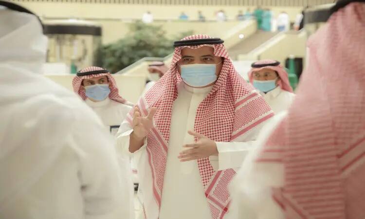 A group of men dressed in traditional Saudi clothing, including a shemagh or ghutra and aqal, as well as the Saudi bisht, and wearing outer masks, during a remembrance. In the center of the group, an attendee is seen using his hand while speaking. Some of the steps and green spaces inside Qassim University can be seen.