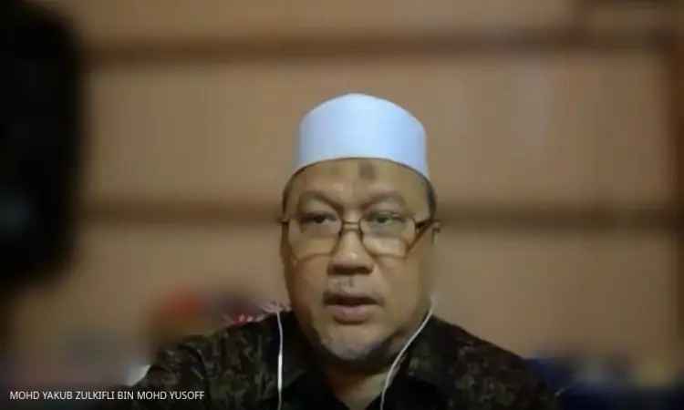 يرتدي الرجل نظارة طبية حديثة، وغترة سعودية بيضاء مع غطاء، ويضع سماعات الأذن، أثناء مكالمة فيديو حول المراقبة. يظهر في الزاوية السفلى رخصة من الشاشة نصية باسم: MOHD YAKUB ZULKIFLI BIN MOHD YUSOFF. الخلفية ضبابية ناعم، مما يضفي مسؤولاً رسمياً يتبع قاً لجامعة القصيم.