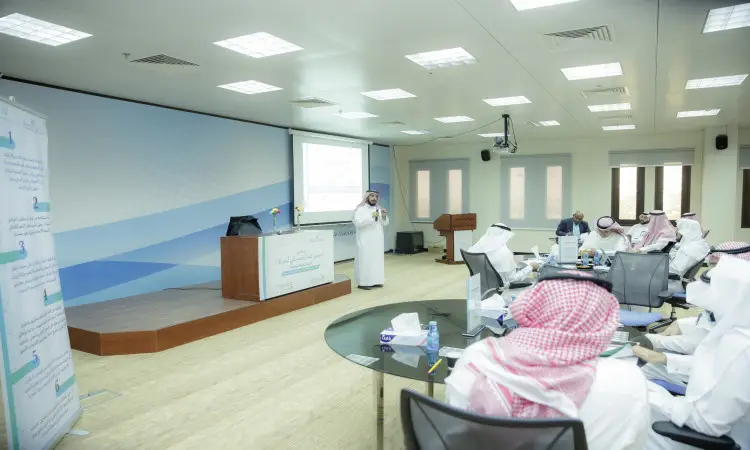 رجل يرتدي الغترة والعقال والبشت السعودي مقدمًا عرضًا حول التنمية في قاعة المؤتمرات الحديثة. يجلس عدد من الحضور بلباسهم الرسمي المكون من الشماغ أو الغترة السعودية والشماع والعقال حول طاولات واسعة، يستمعون باهتمام ويسجلون الملاحظات، بينما يُعرض مراقبة العرض على شاشة العرض أمامهم.